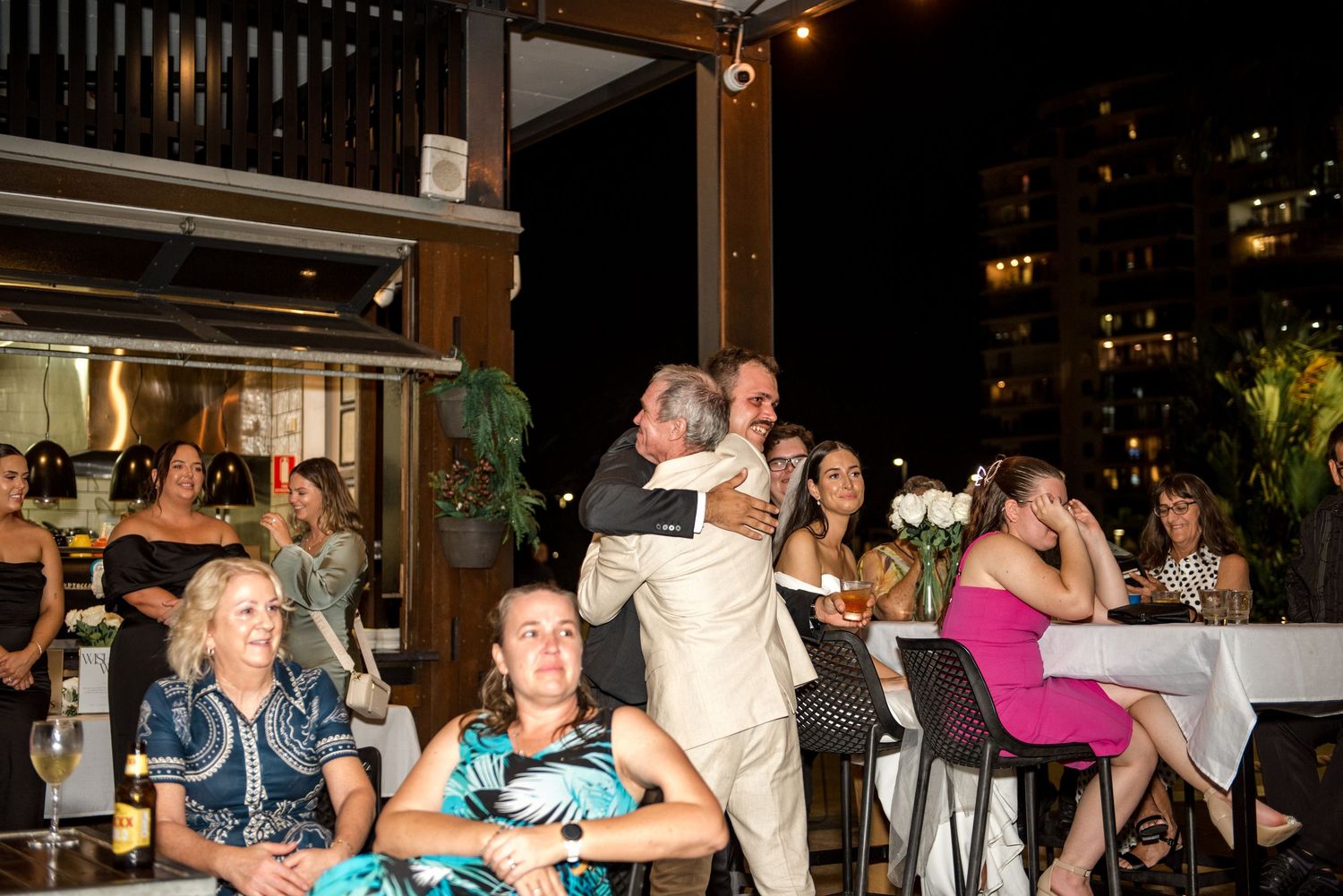 Wedding guests watch as two men in suits embrace during an outdoor evening reception celebration.