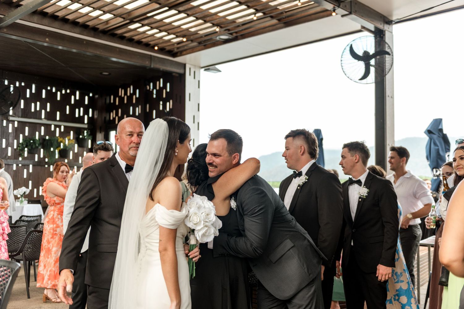 Groom receiving warm embrace from guest while bride stands nearby at wedding reception.