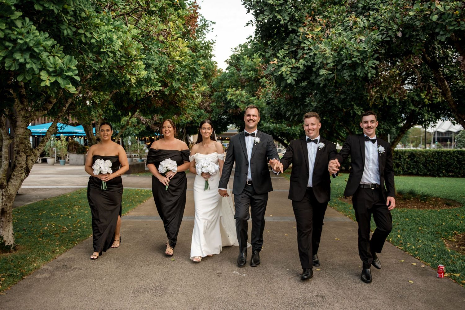 Wedding party in black and white walking through a tree-lined park path.