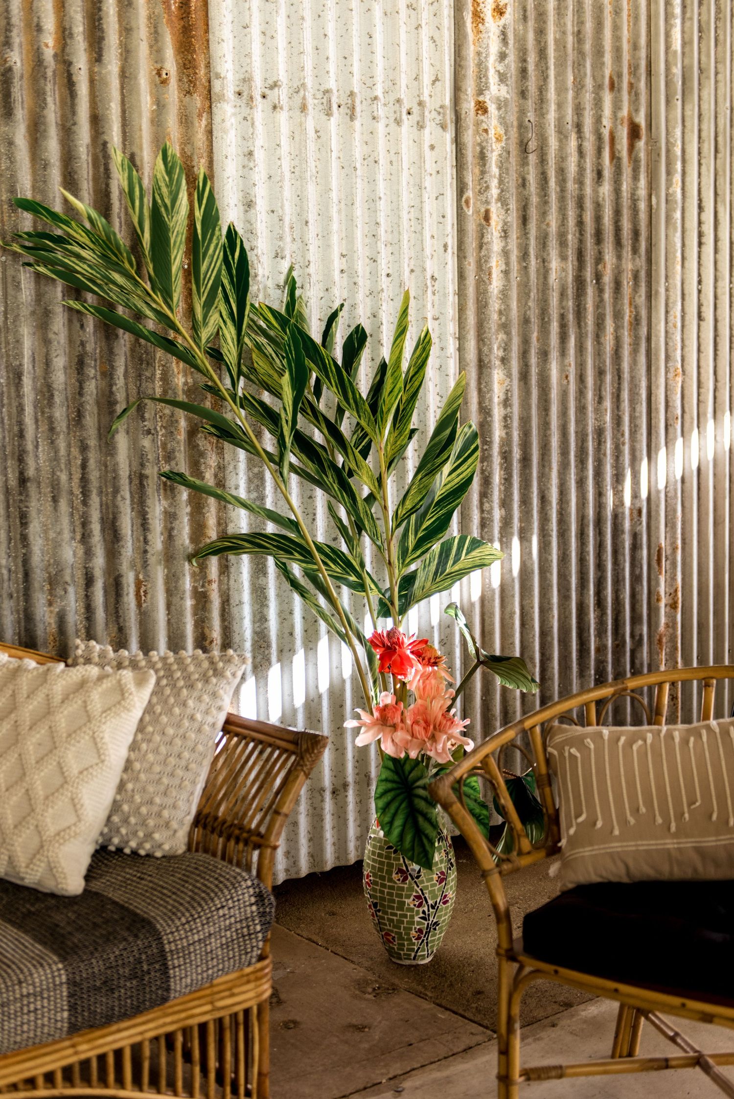 Tall tropical floral arrangement in mosaic vase beside rattan furniture against rustic corrugated iron wall.