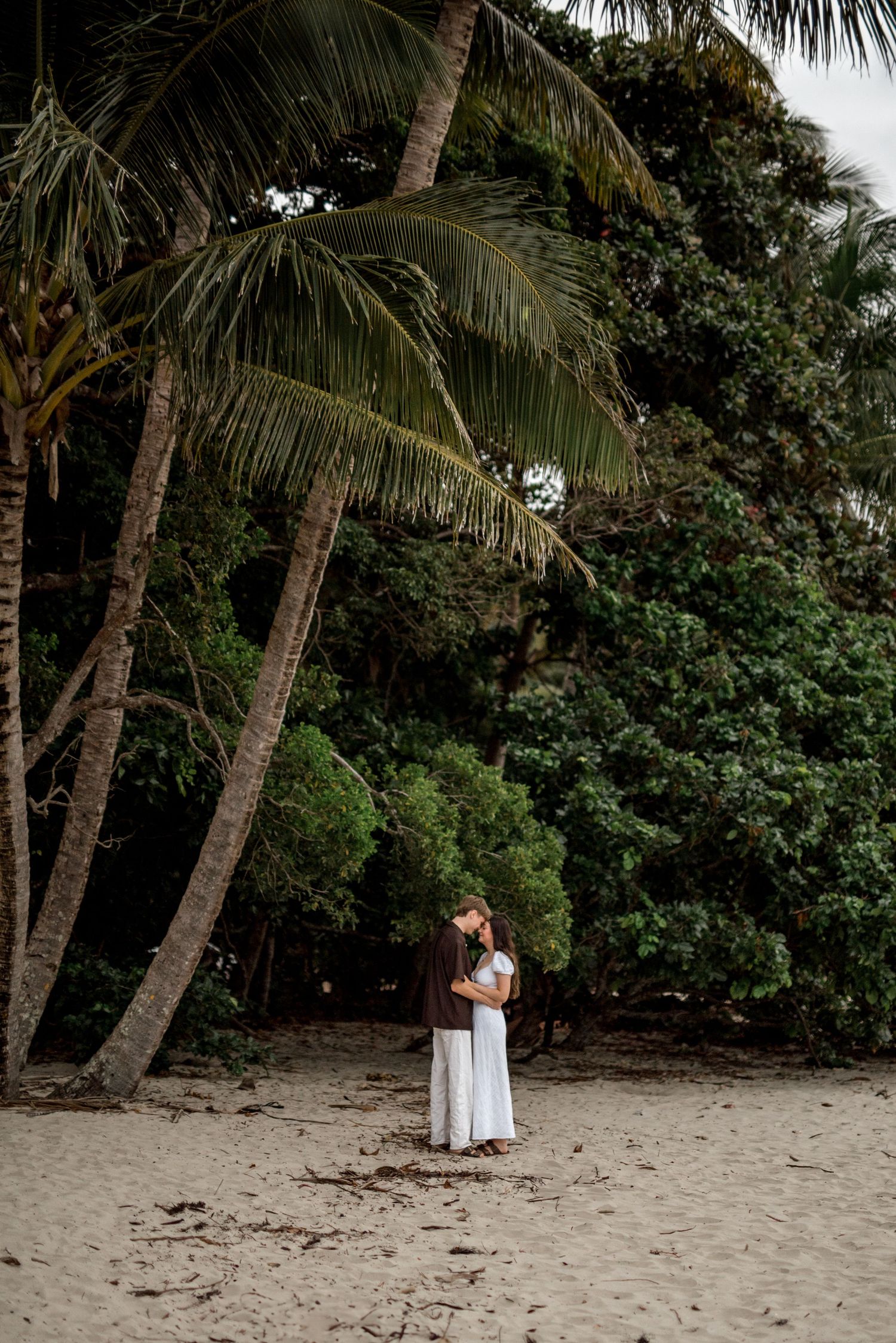 Couple embracing and kissing under tall palm trees on a sandy tropical beach.