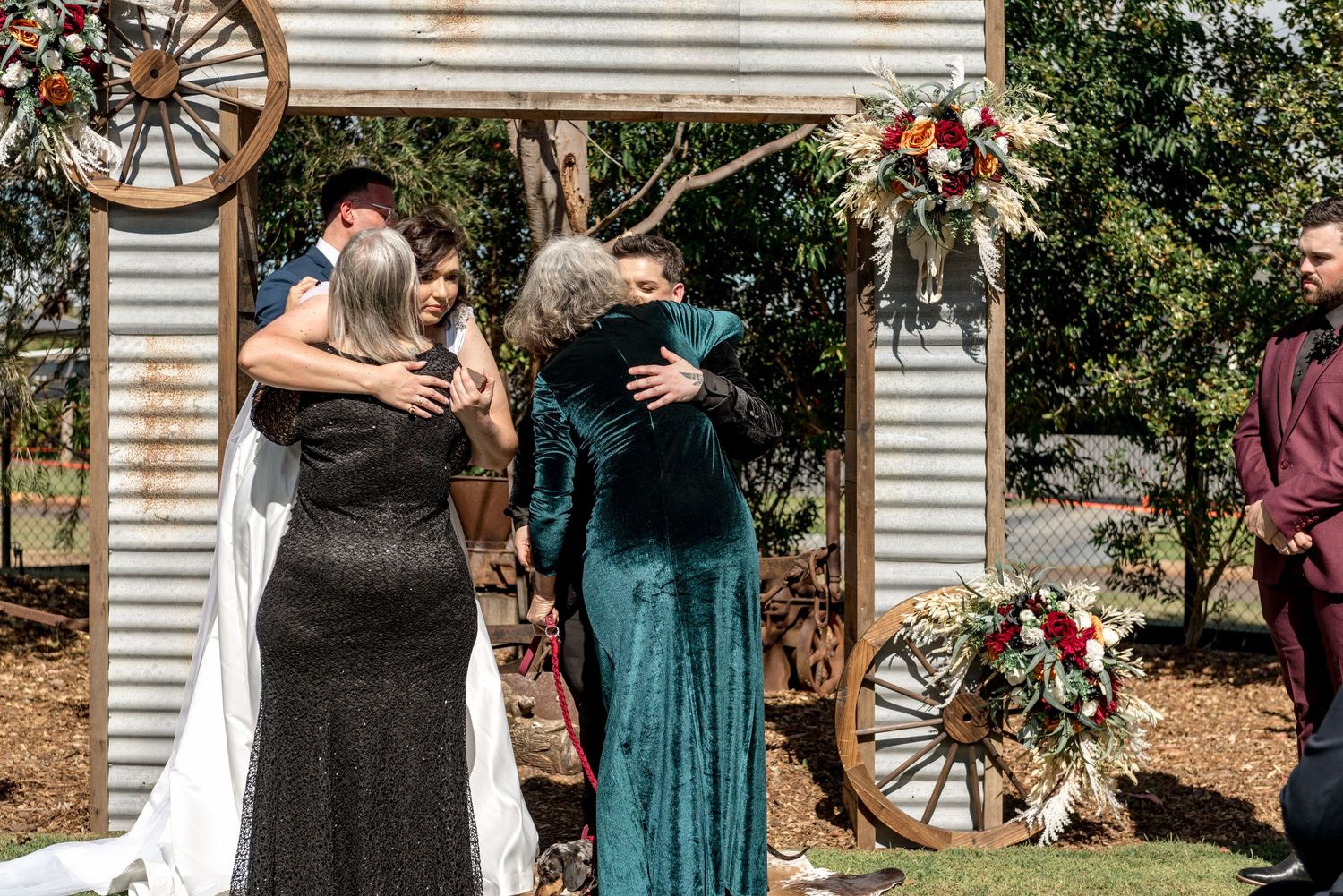 Emotional wedding guests embrace near a rustic corrugated iron arch decorated with flowers.