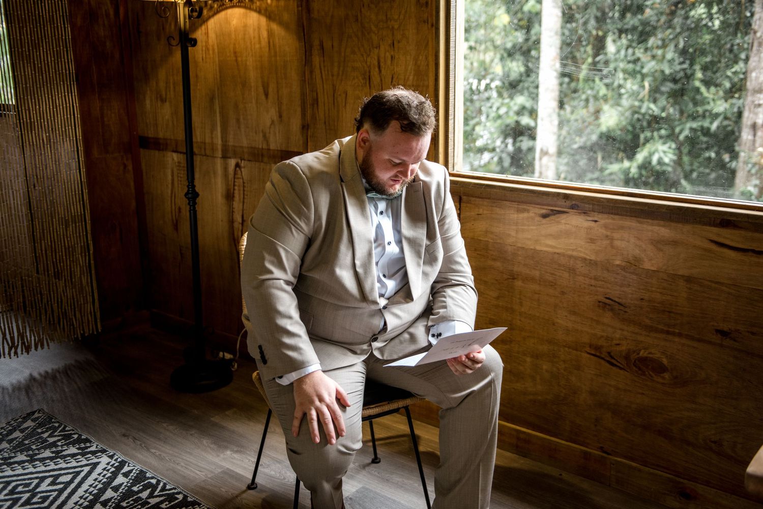Groom in tan suit reading note while seated in rustic wooden room with natural light.