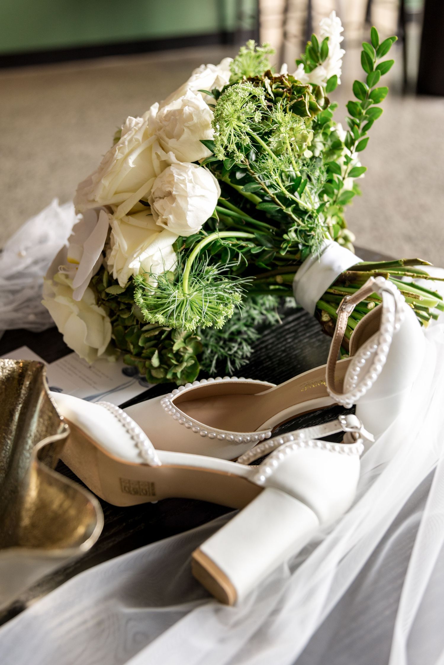 Bridal flat lay featuring white floral bouquet, pearl-strap heels, and flowing veil on dark surface.