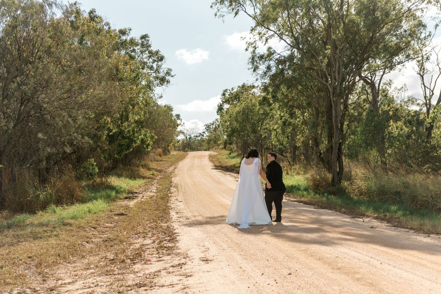 Bride and groom walk hand-in-hand away from camera down a sandy rural dirt road lined with trees.