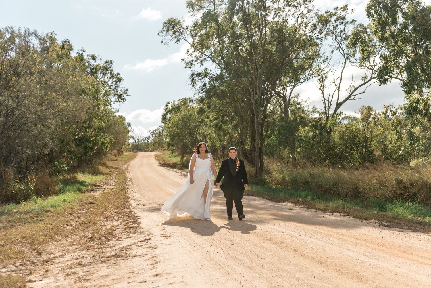 Bride in white gown and groom in black suit walk toward camera on a sunny rural dirt road.