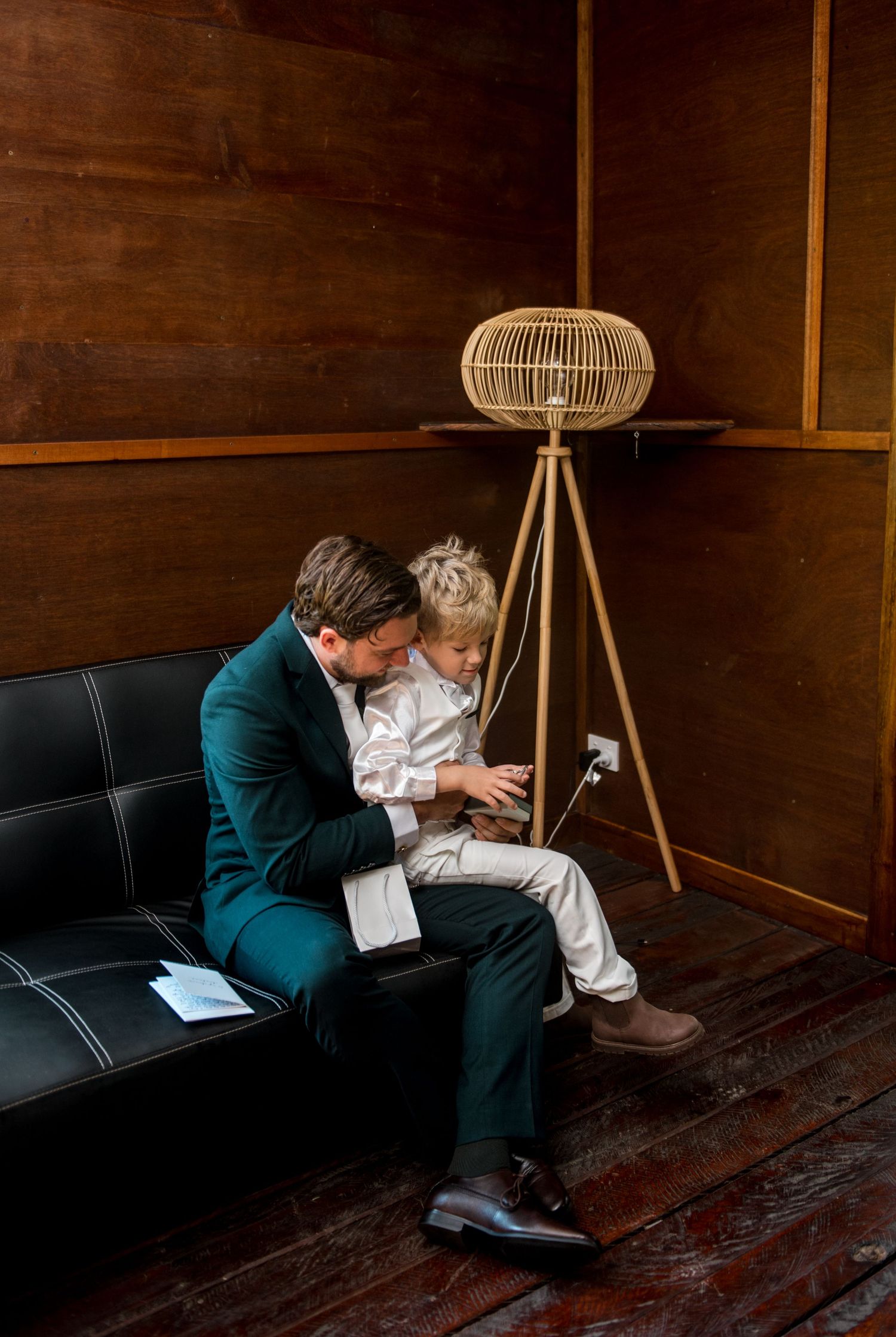 Groom in teal suit sitting with young boy on leather couch before wedding.
