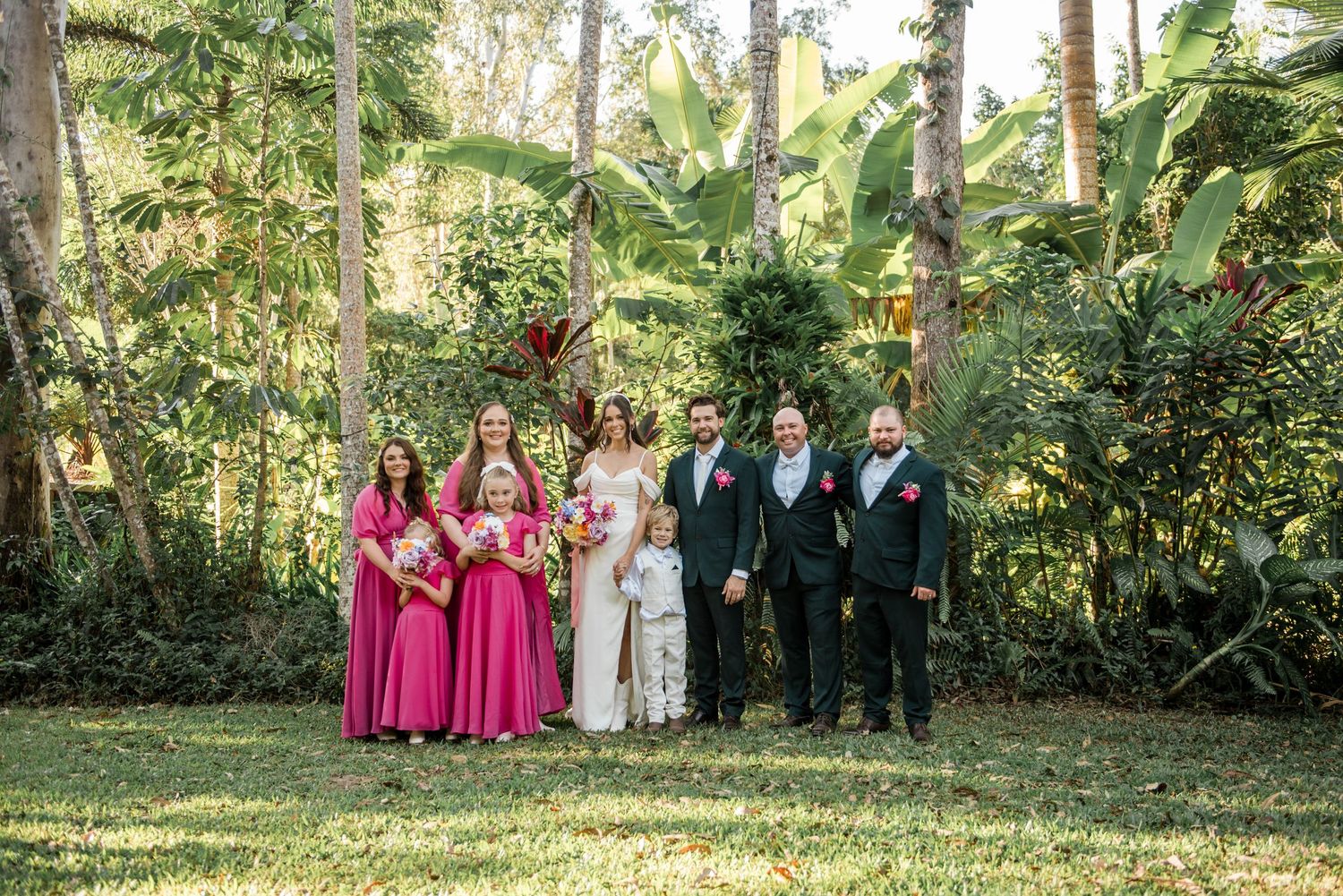 Wedding party posing in tropical garden with bridesmaids in hot pink gowns and groomsmen in grey suits with pink florals.
