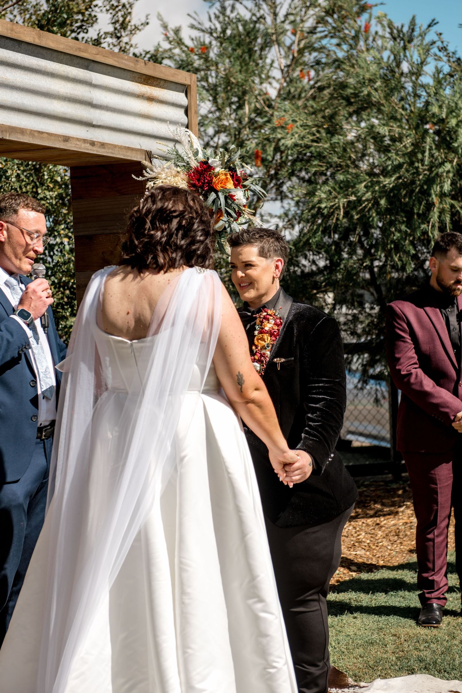Bride in white gown with veil holds hands with groom in black velvet suit at outdoor rustic wedding ceremony.