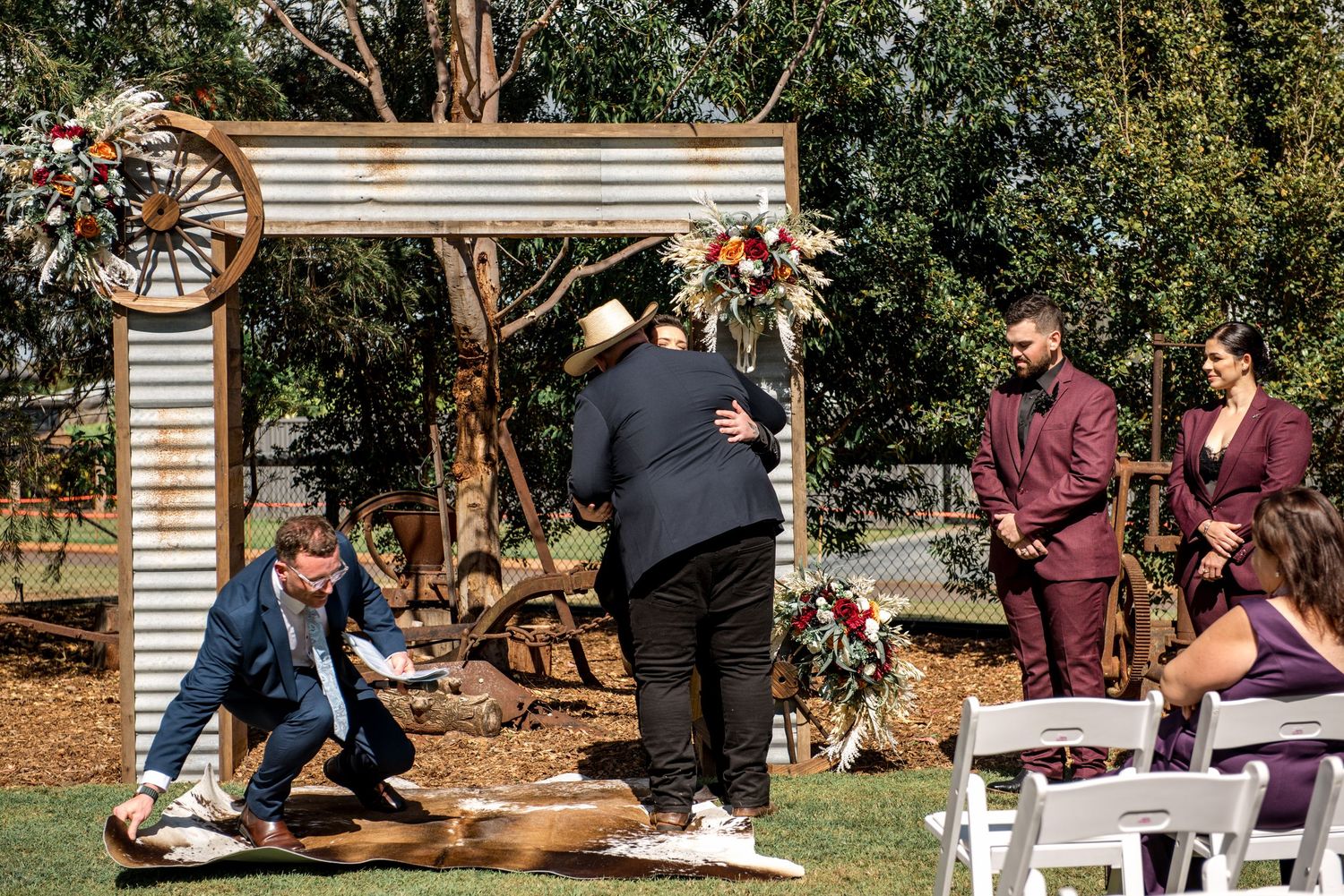 Groom greets guest at rustic outdoor wedding altar decorated with dried florals and corrugated iron backdrop.