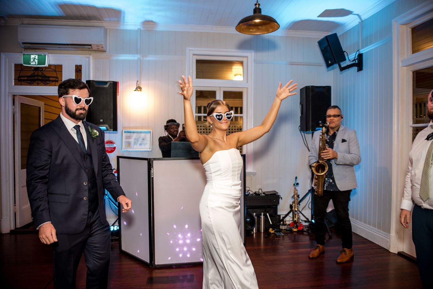 Bride in white gown with novelty sunglasses dancing with groom while a saxophonist performs at wedding reception.