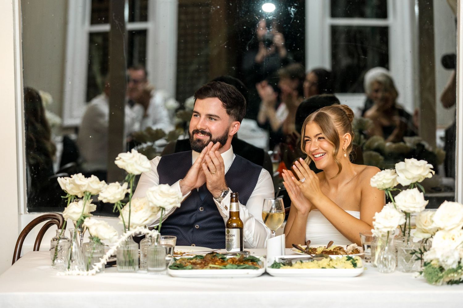 Bride and groom clapping and smiling together at their elegant wedding reception dinner table.