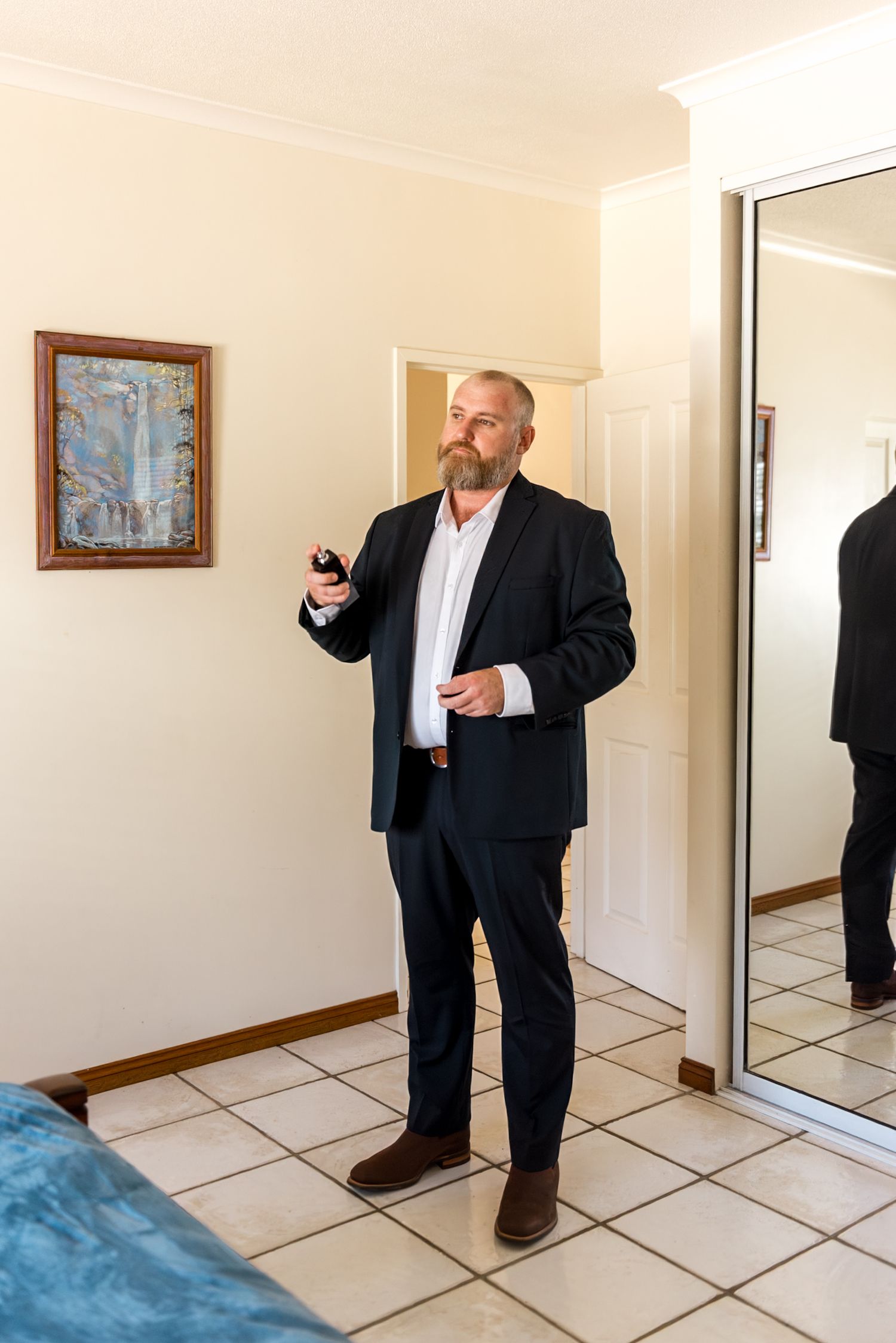 Bearded groom in navy suit holding cologne bottle while getting ready in bedroom.