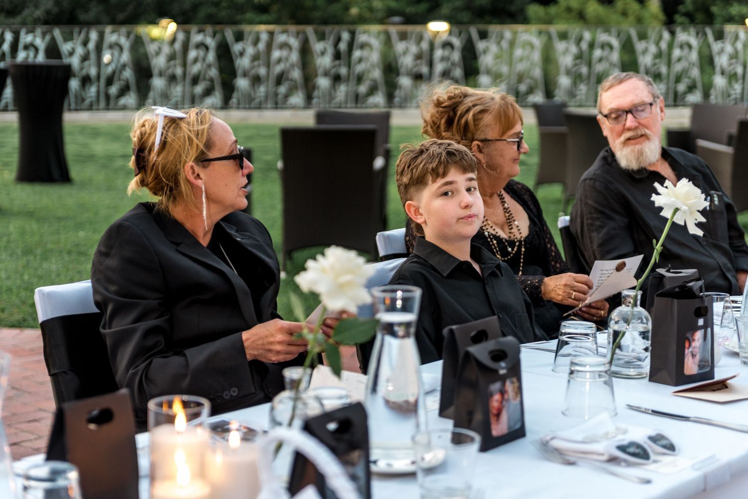 Wedding guests seated at outdoor reception table holding white flowers with candles and floral centerpieces at dusk.