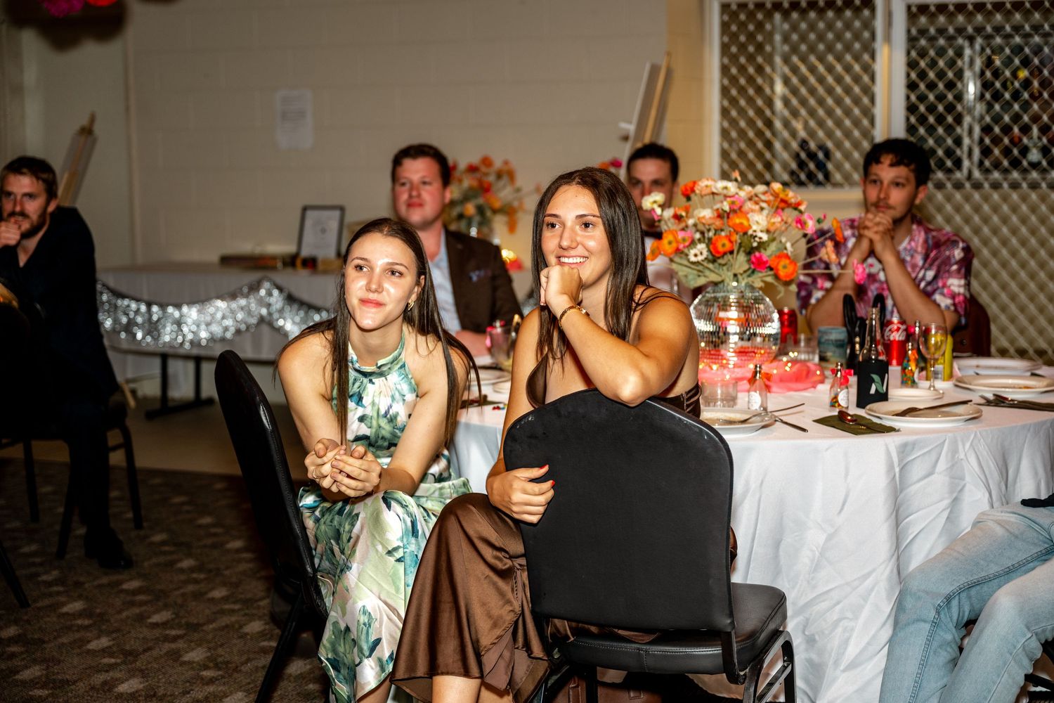 Two smiling women enjoying wedding reception seated at elegantly decorated floral table.