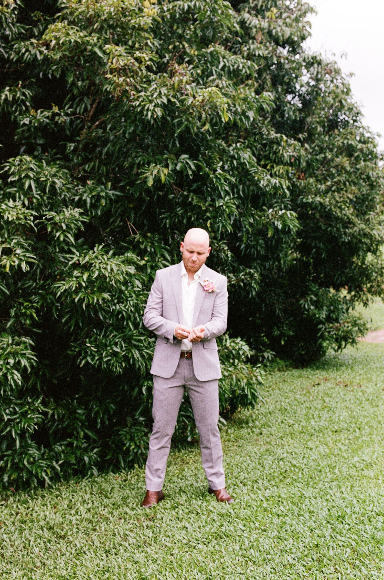 Bald groom in light gray suit with floral boutonniere standing on green grass in front of trees.