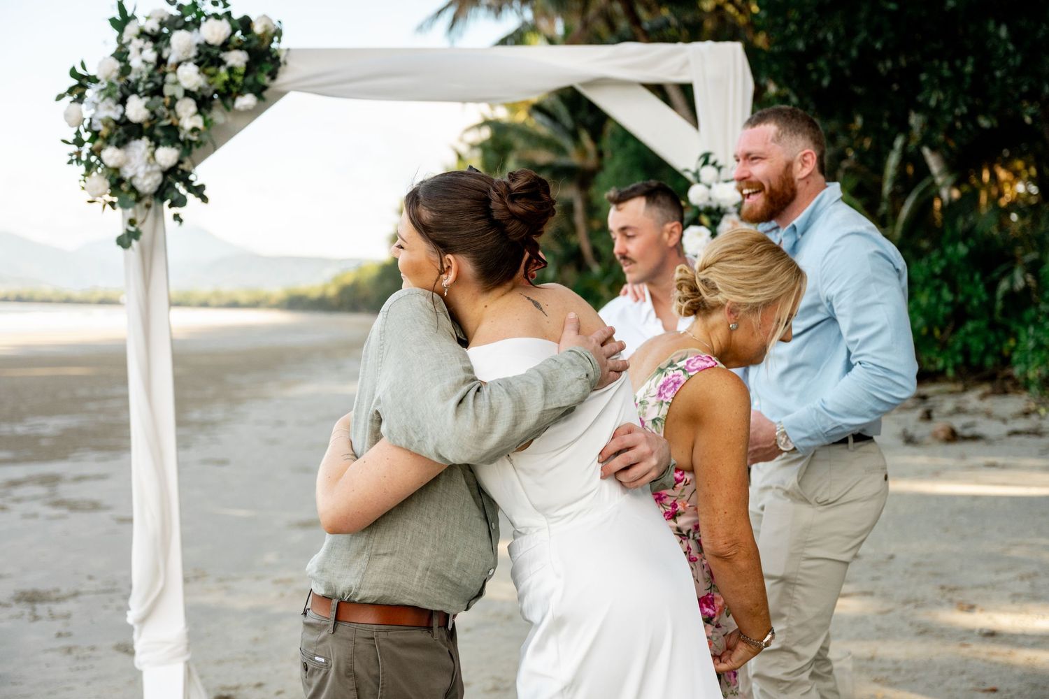 Wedding party exchanging hugs after beach ceremony under a white floral arch with palm trees at Four Mile Beach, Port Douglas.
