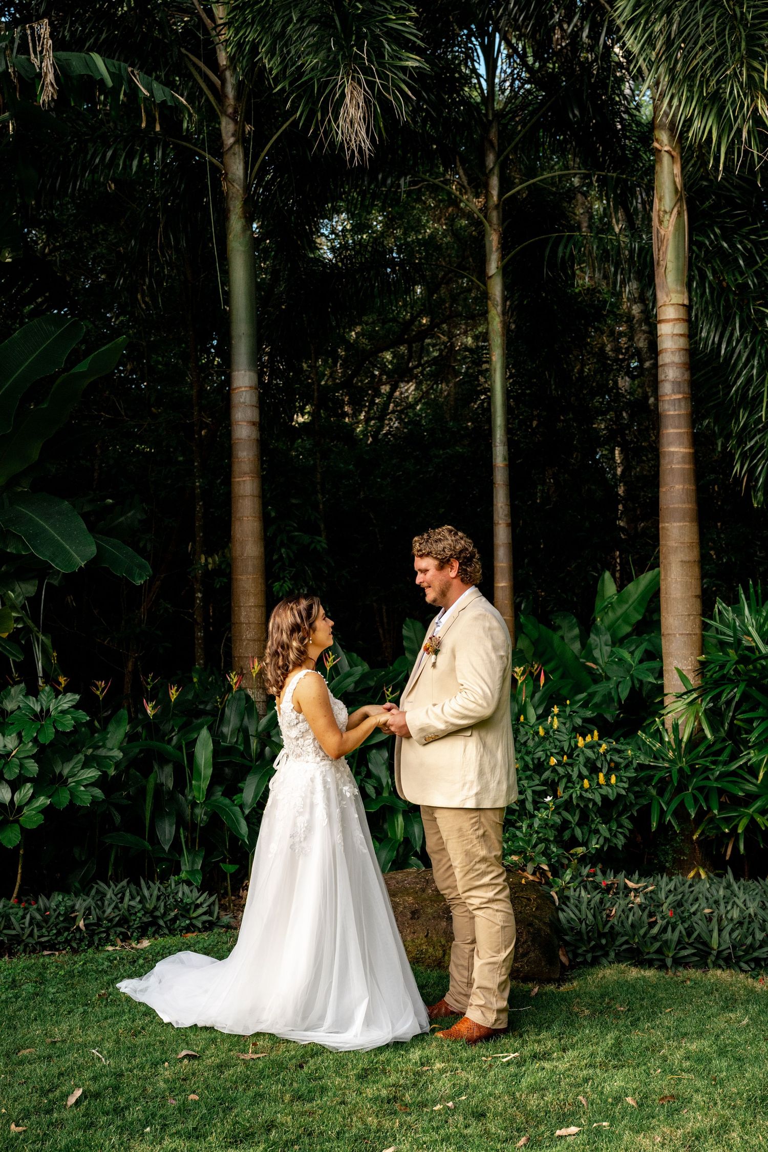 Smiling bride and groom hold hands facing each other in sunlit tropical garden after wedding ceremony.