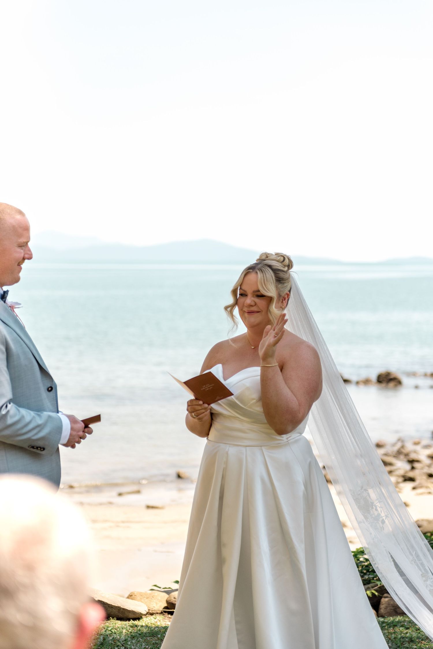 Emotional bride reads vows from book during beachside ceremony with ocean and mountains behind her.