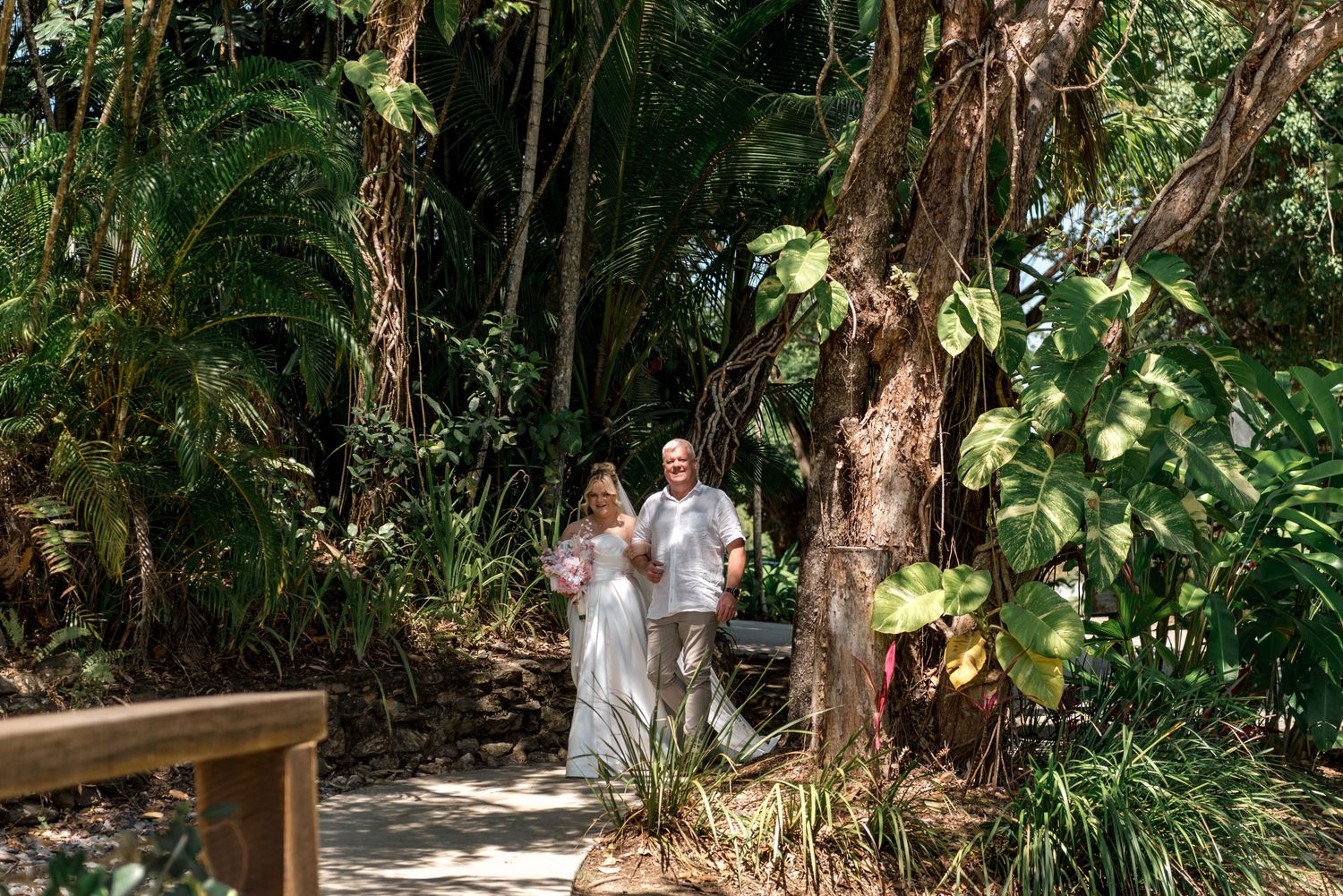 Bride and groom walk hand in hand through lush tropical garden pathway on wedding day.