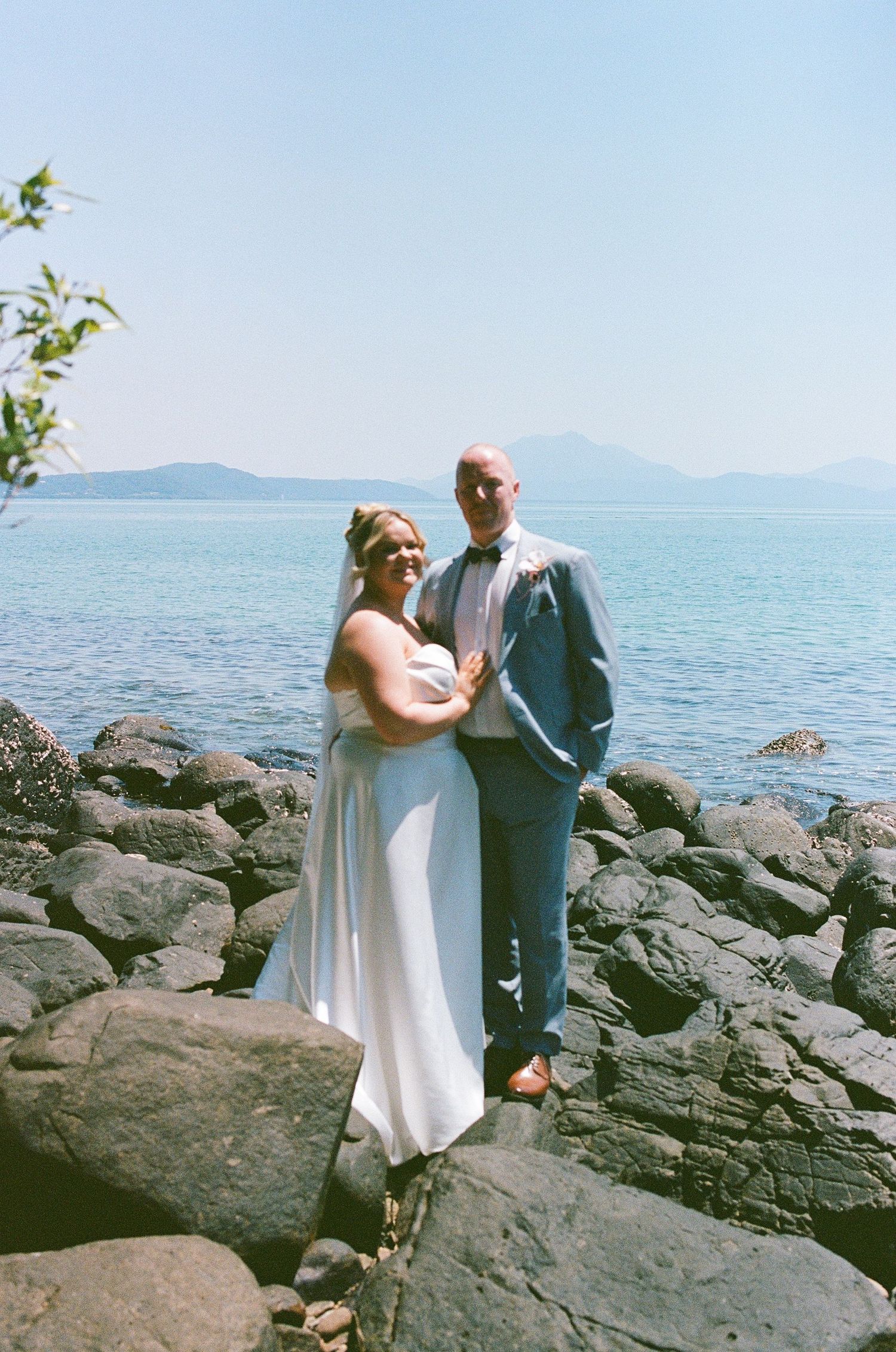 Bride and groom stand on rocky shoreline with turquoise ocean and mountain views behind them.