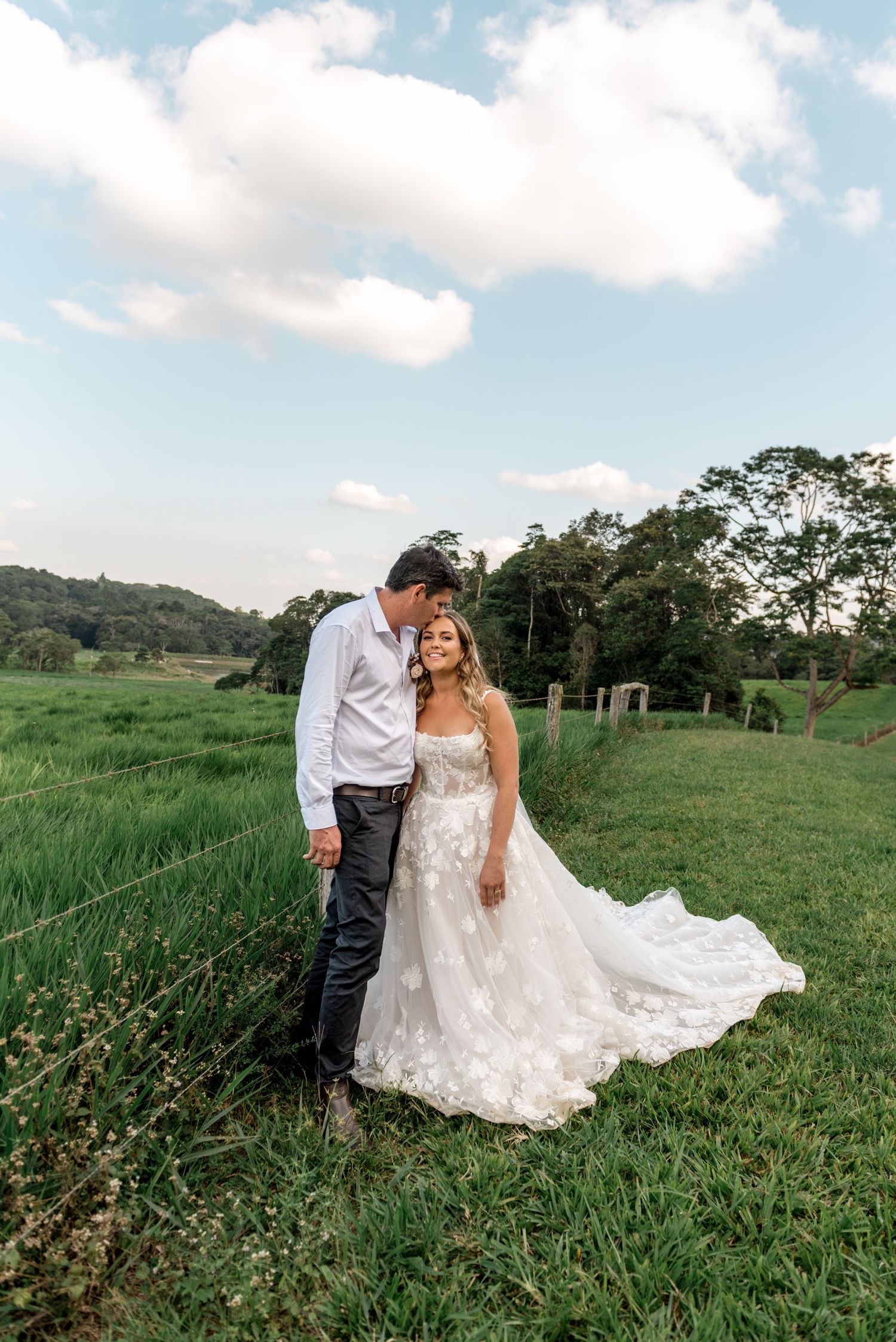 Groom tenderly kissing bride on the cheek in a lush green field under a partly cloudy sky.
