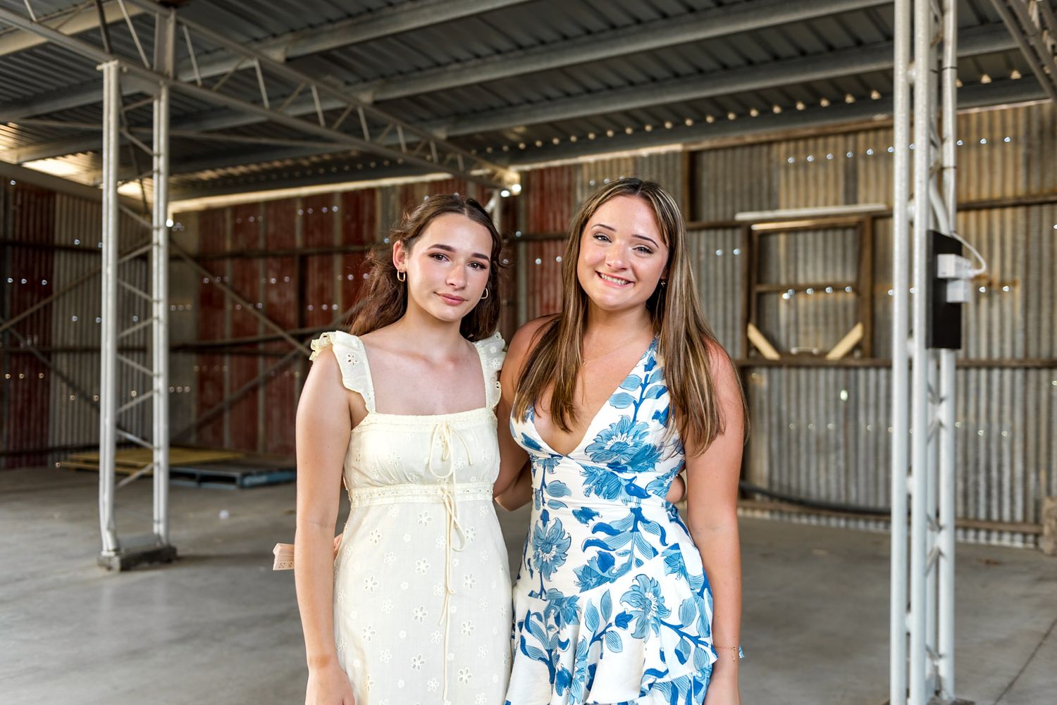 Two young women in summer dresses pose together smiling inside a rustic barn wedding venue.