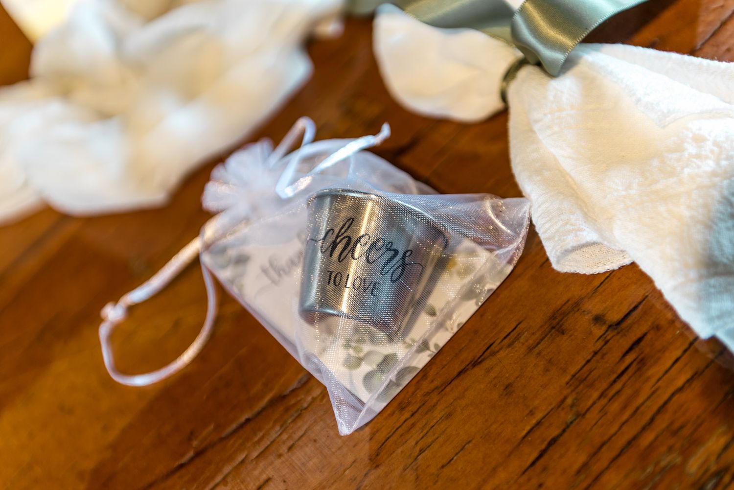 Silver cheers to love shot glass wedding favor in sheer organza bag on wooden surface.