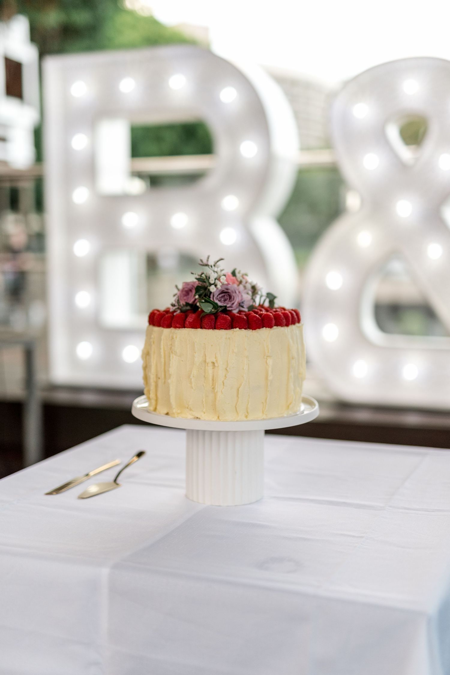 Elegant single-tier white wedding cake topped with raspberries and pink flowers on white cake stand.