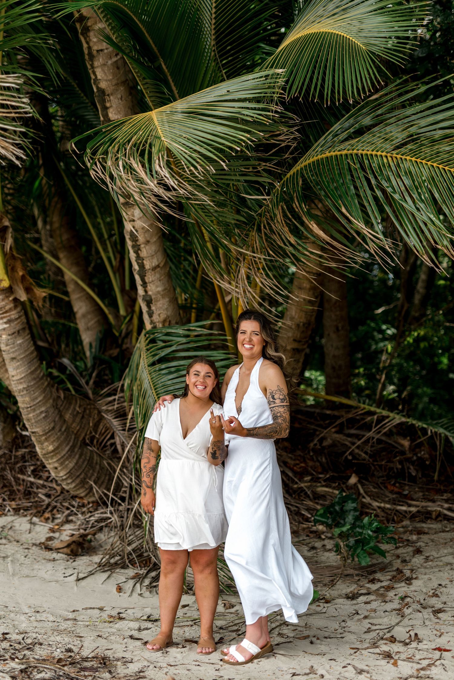 Two women in matching white dresses smiling together on a sandy beach with lush palm trees behind them.