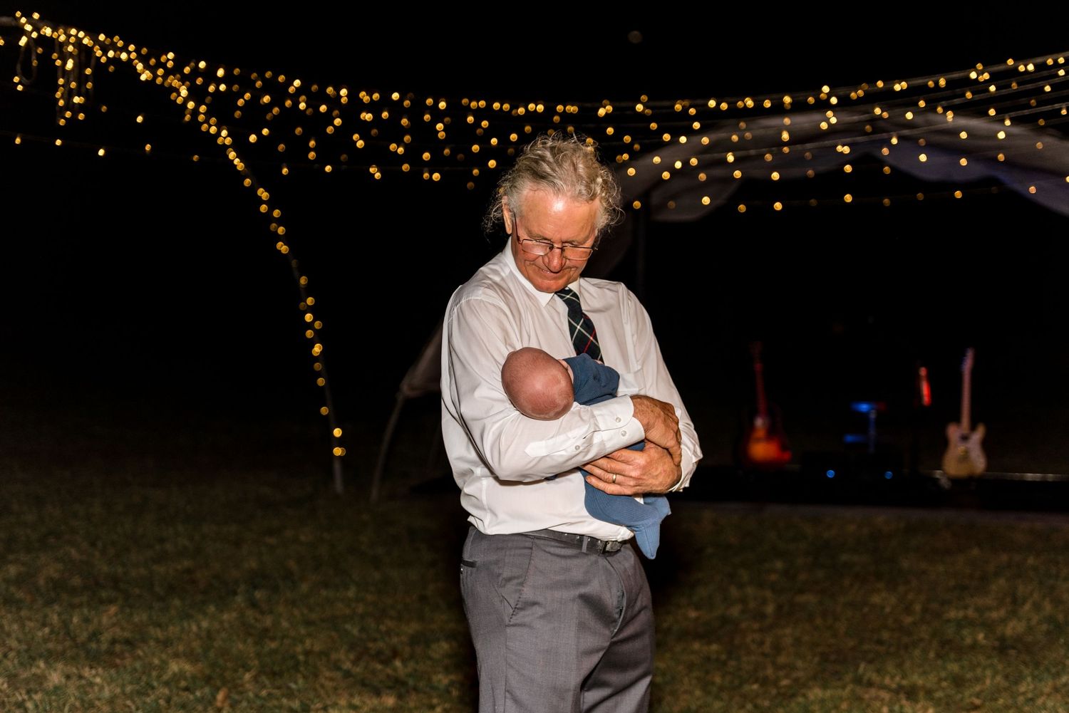 An elderly man in a suit tenderly holds a newborn baby under twinkling string lights at an outdoor evening event.