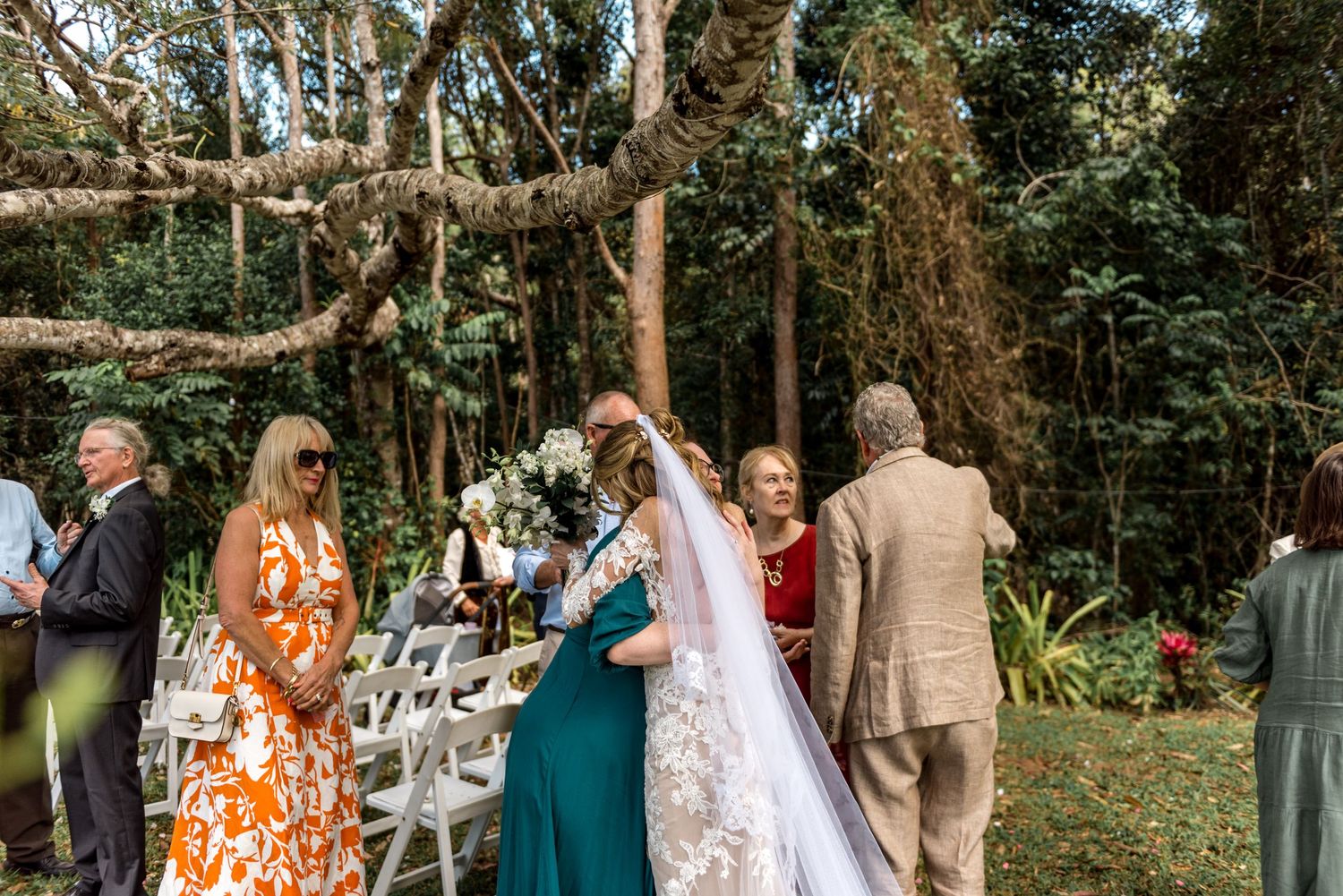Bride in lace gown greeted by guests at outdoor garden wedding ceremony.