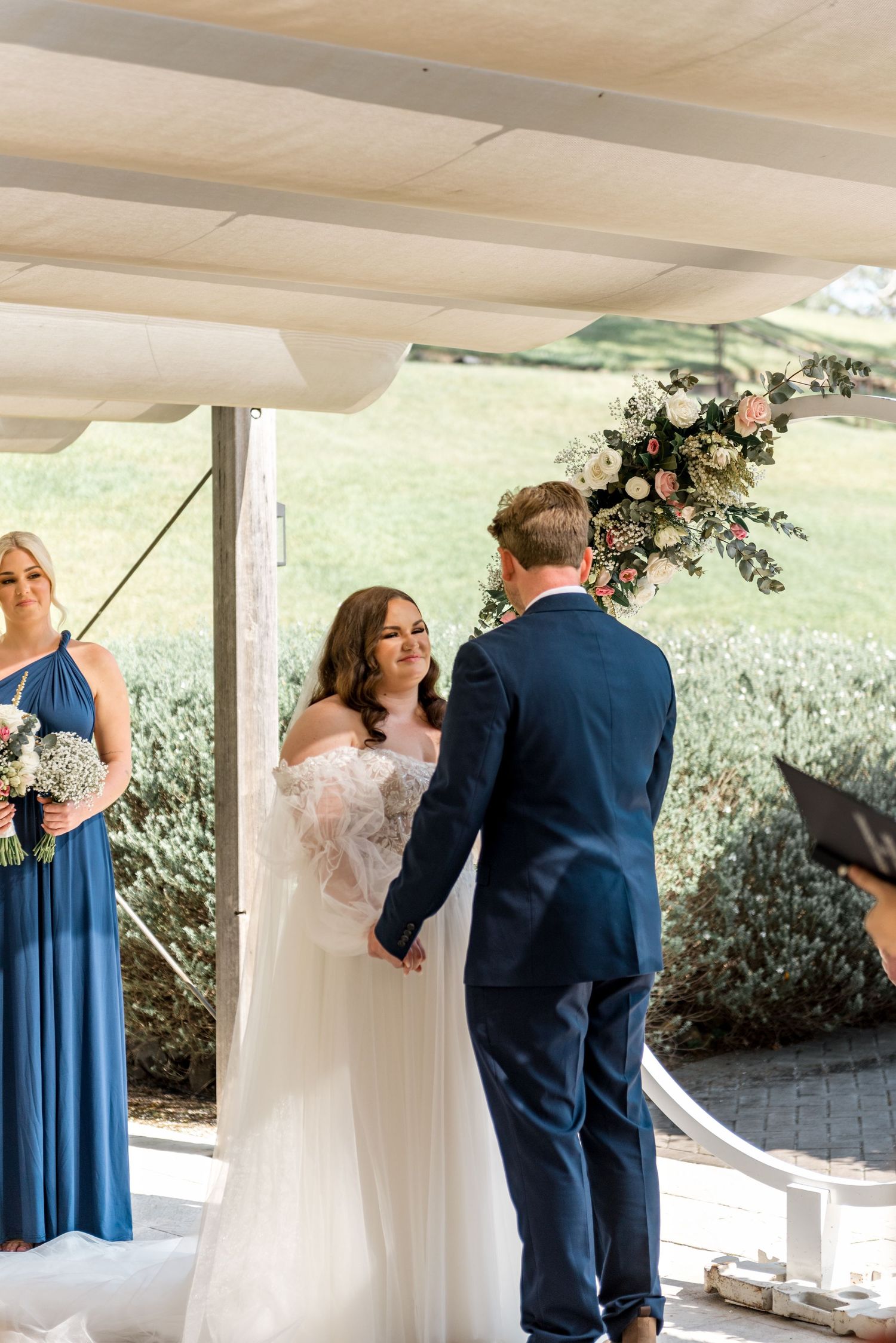 Bride and groom hold hands at the altar with a floral arch during outdoor wedding ceremony.