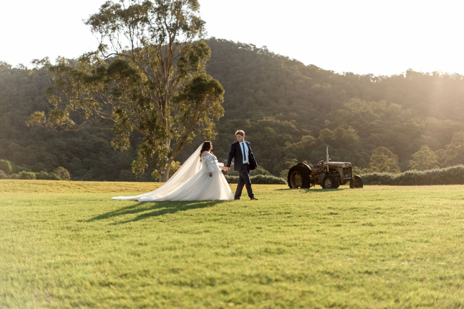 Bride and groom walk across a sunlit field with a vintage tractor and rolling hills behind.
