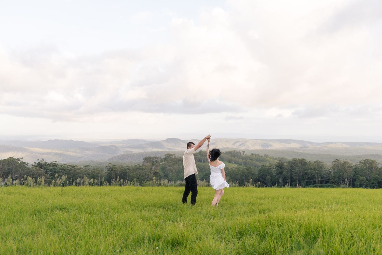 Couple dancing joyfully in a lush green field with rolling hills and cloudy sky backdrop.