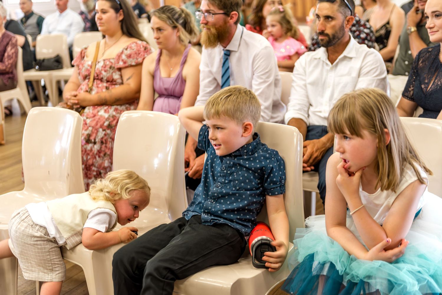 Children and adults seated at wedding ceremony, with young boy holding a drink among the guests.