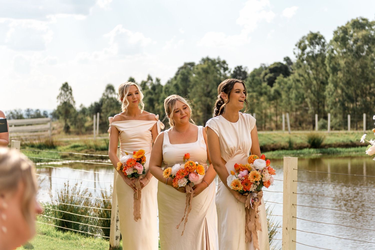 Three bridesmaids in cream dresses holding vibrant orange and white bouquets beside a scenic pond at a wedding.