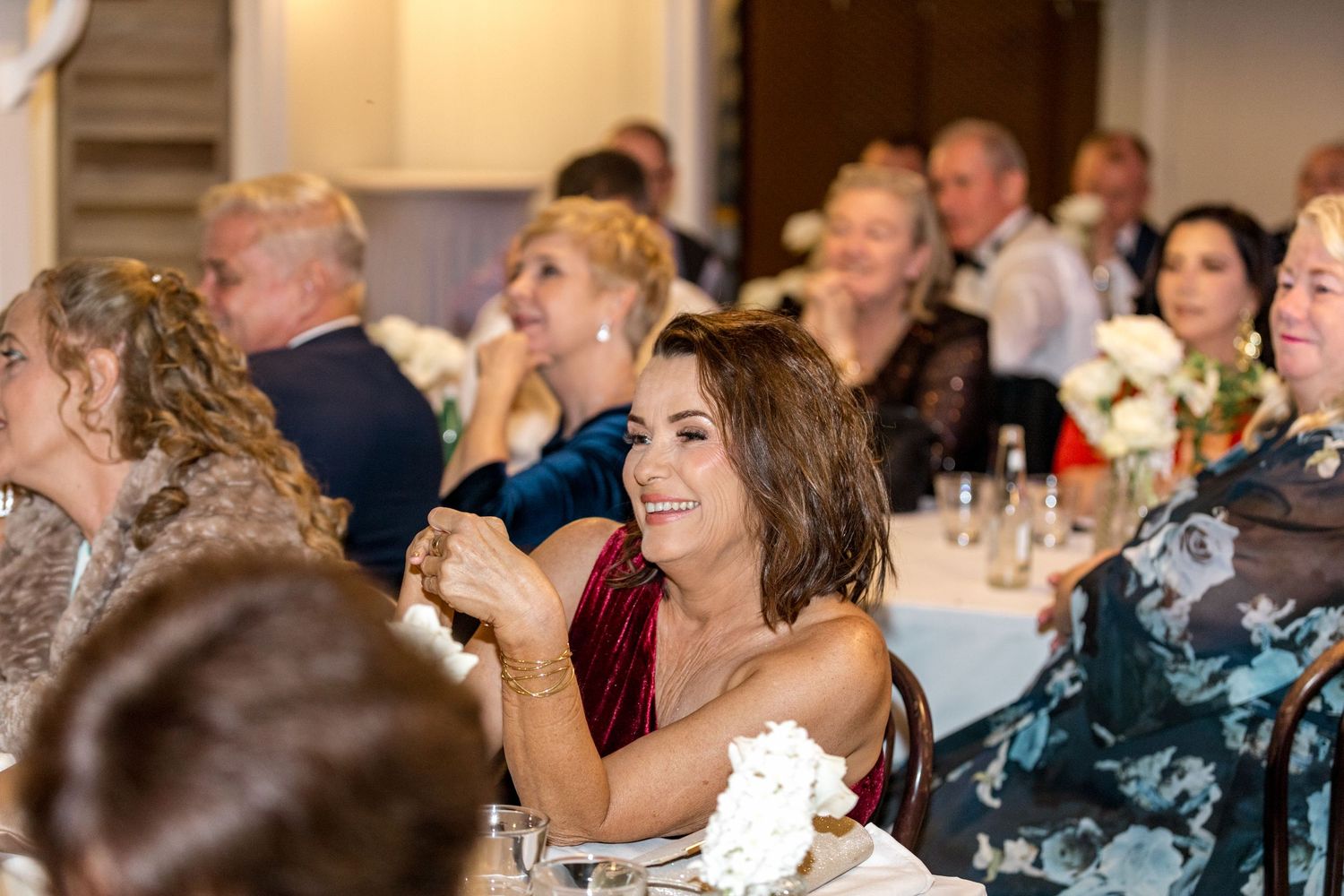 Guests smiling and clapping during speeches at an elegant wedding reception with floral decor.