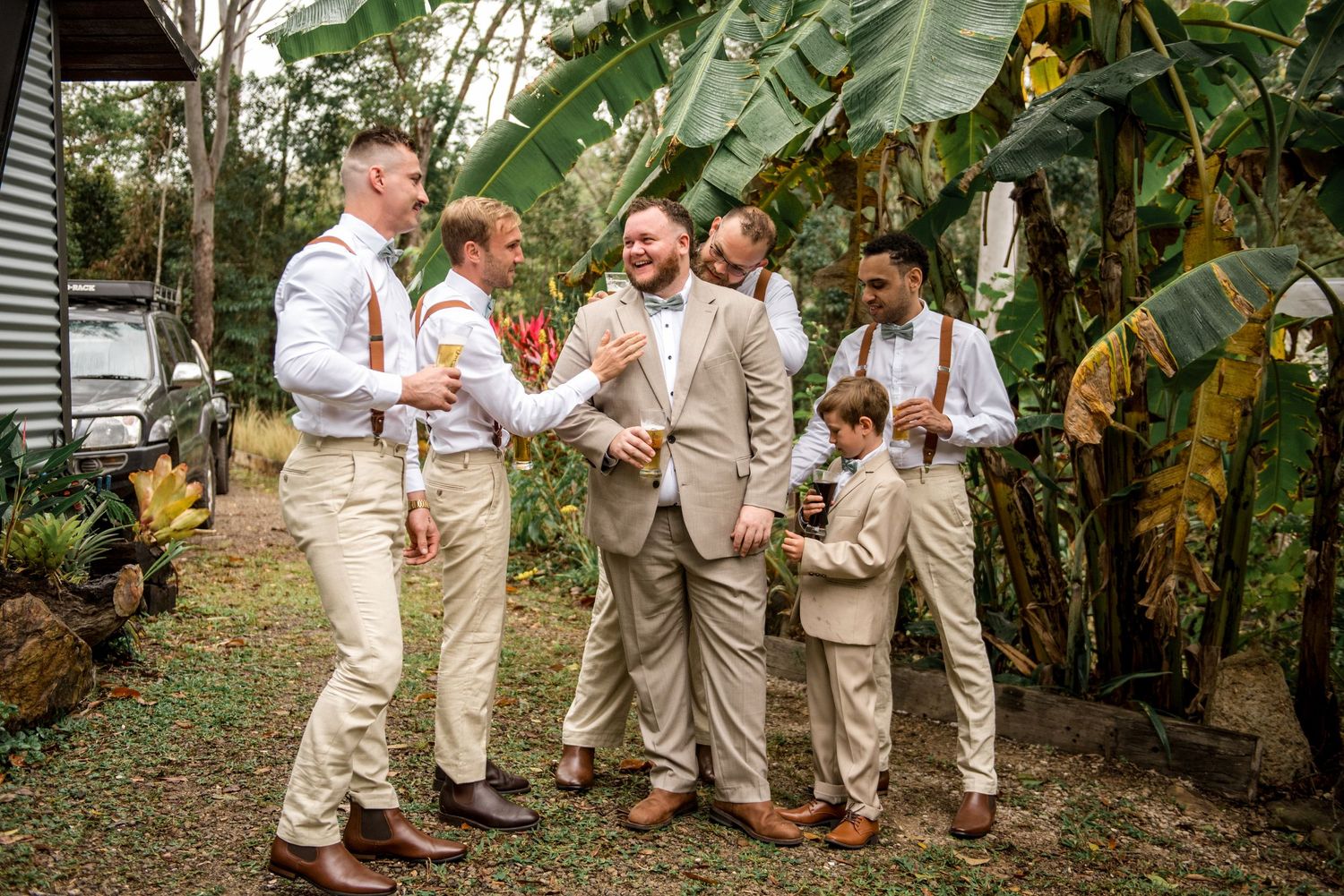 Groomsmen in tan suits laughing together outdoors surrounded by tropical greenery before a wedding ceremony.