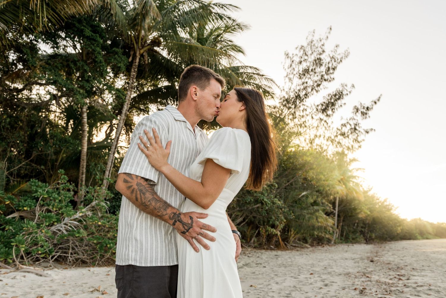 Couple kissing on a sandy beach at sunset surrounded by lush tropical palm trees and warm golden light.