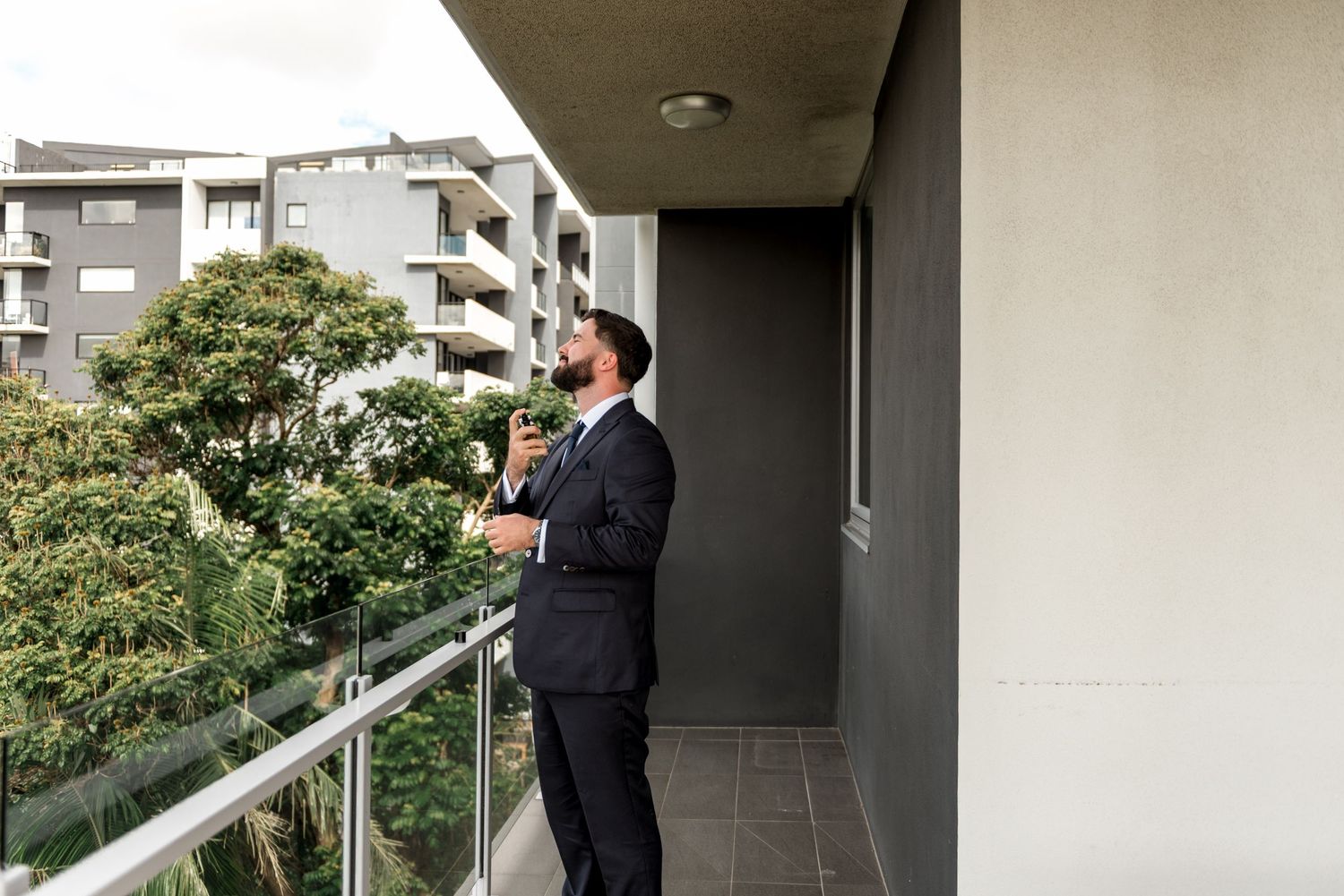 Stylish groom in a navy suit standing on a balcony adjusting his tie with city views.
