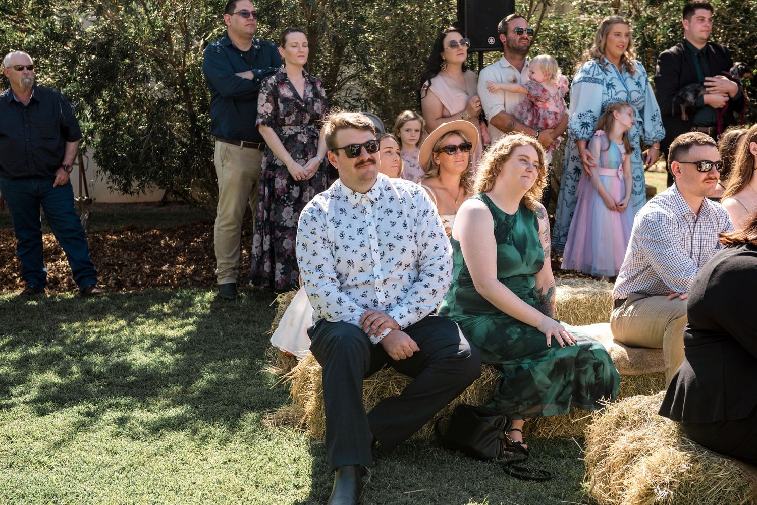 Wedding guests seated on hay bales outdoors, man in floral shirt and woman in teal dress watching ceremony.