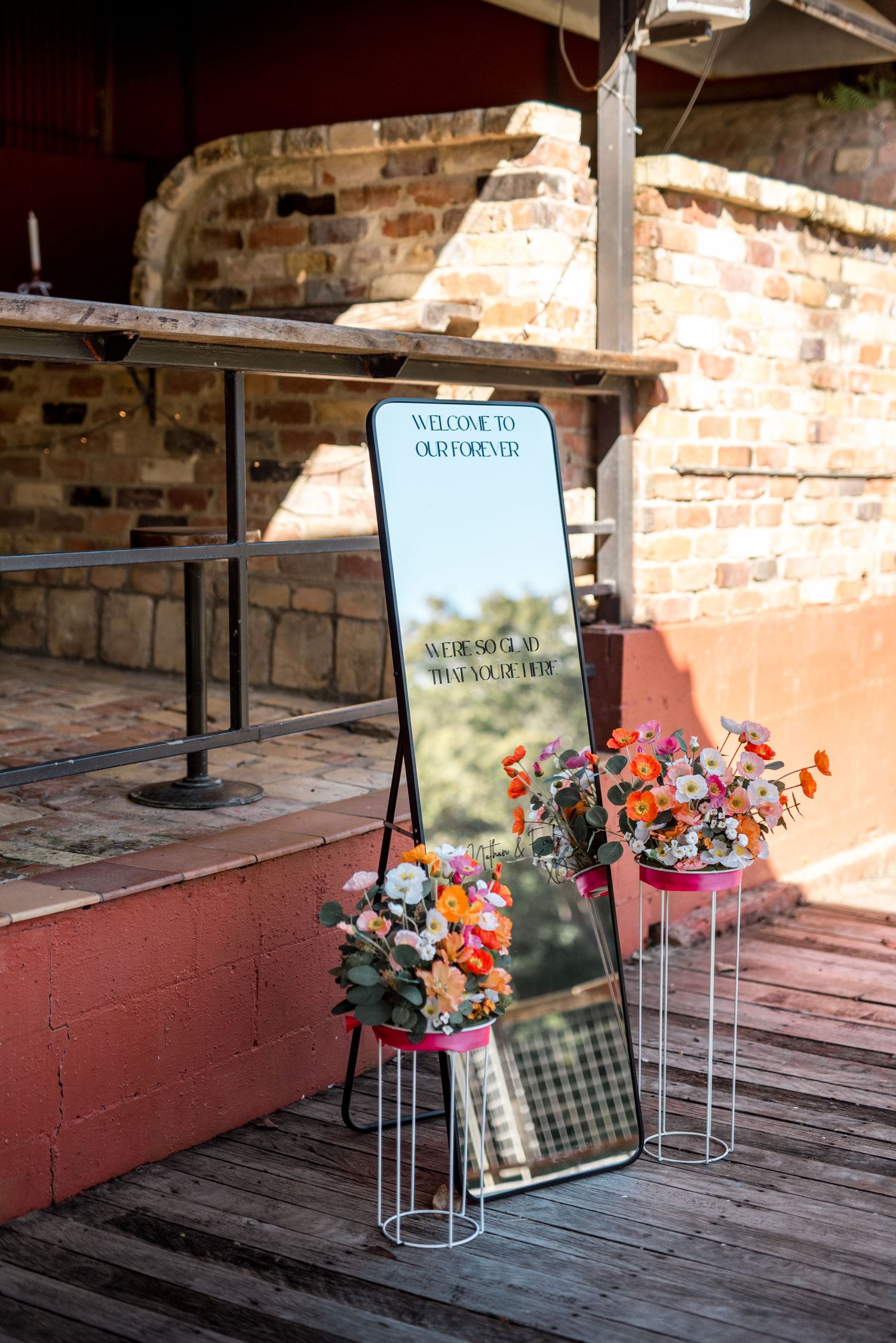 Full-length mirror wedding welcome sign reading Welcome to Our Forever, flanked by colorful floral arrangements on white stands.