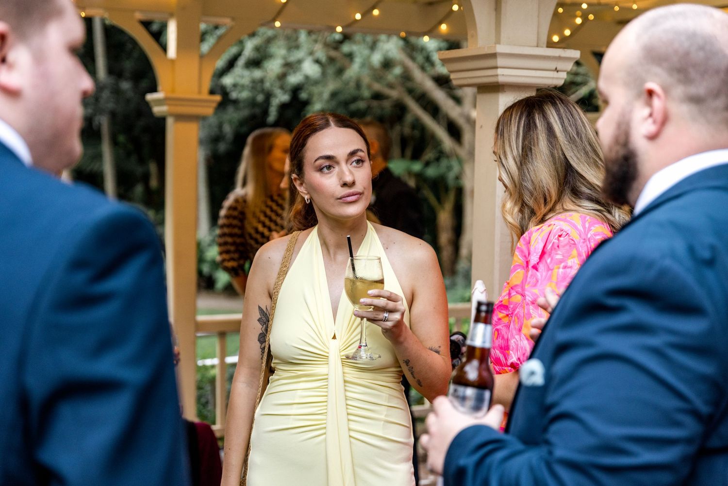 A woman in a yellow halter dress holds a drink while chatting at an outdoor wedding reception.