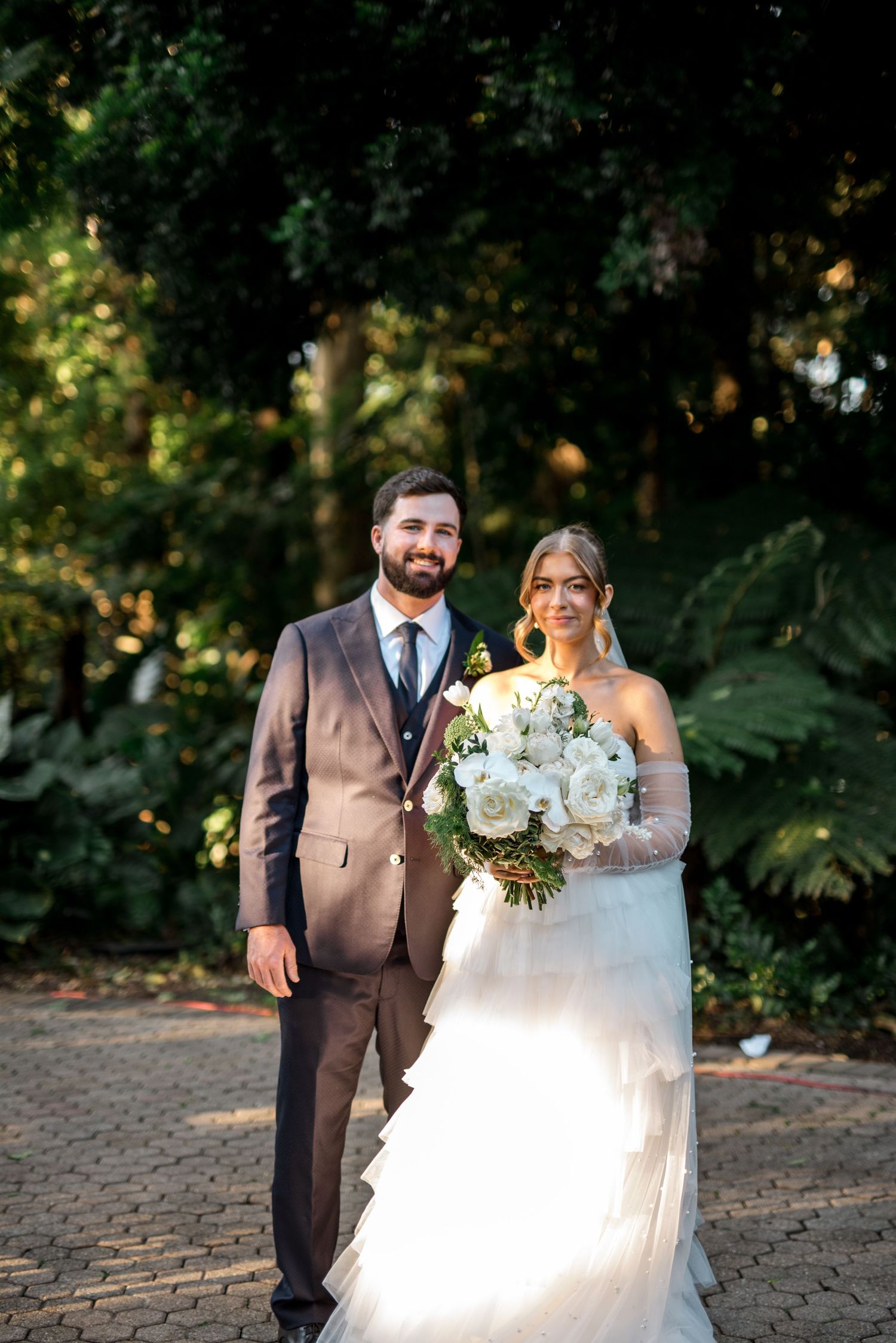 Bride and groom pose together on brick path surrounded by lush green trees after wedding.