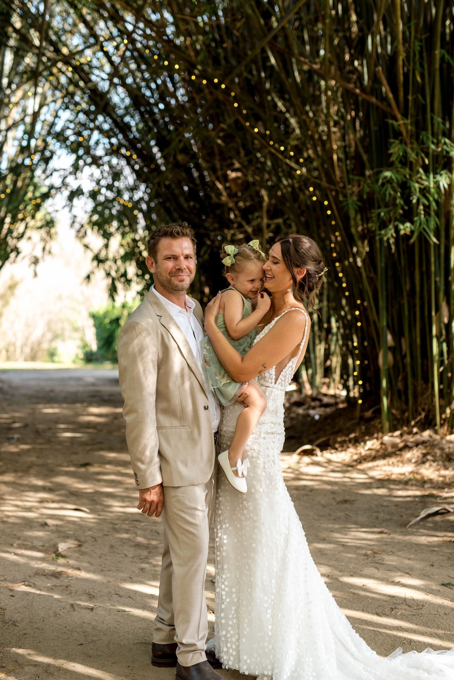 Bride and groom holding young daughter in green dress under bamboo arch with fairy lights at outdoor wedding.