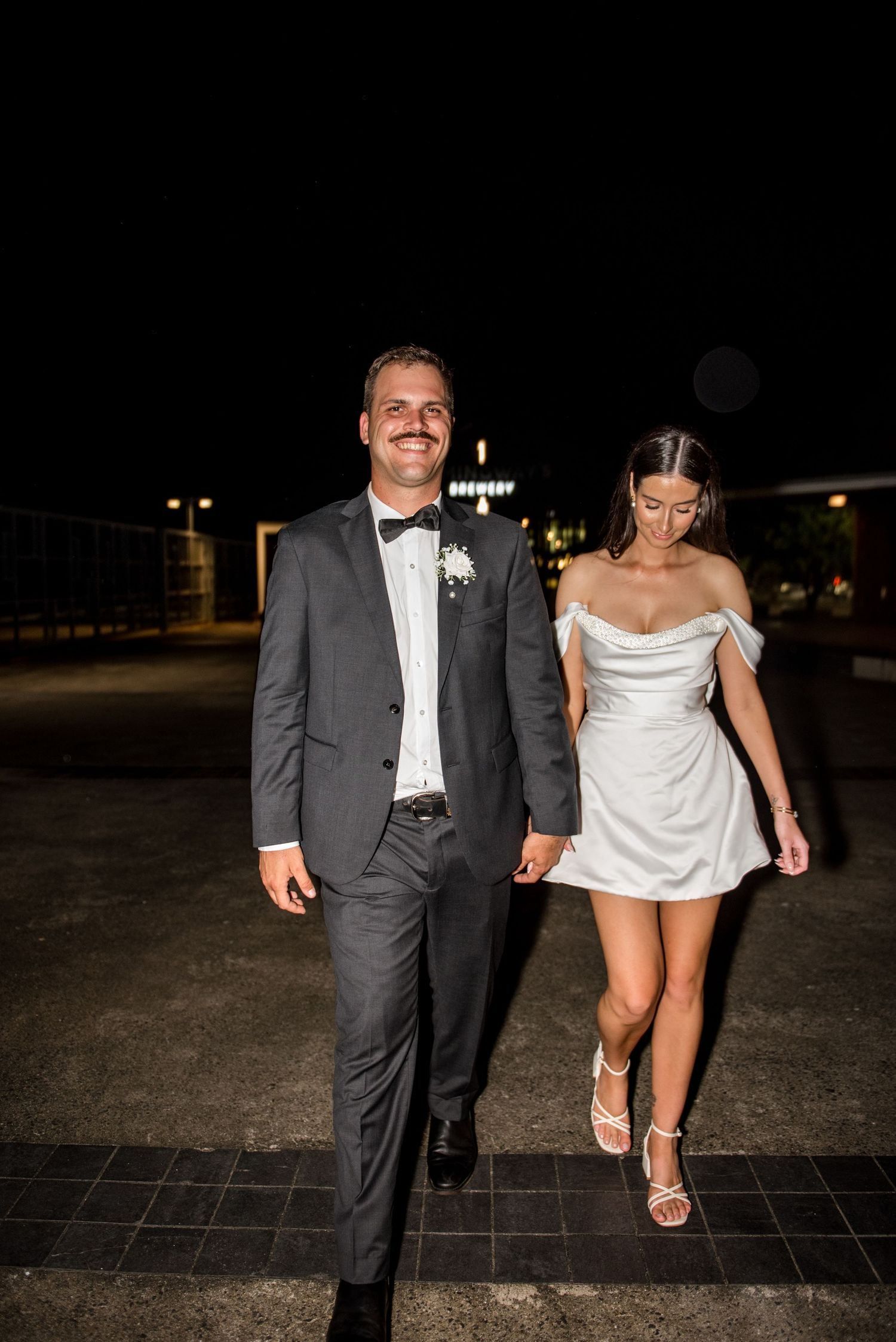 Smiling groom in tuxedo and bride in white mini dress walk hand in hand at night.