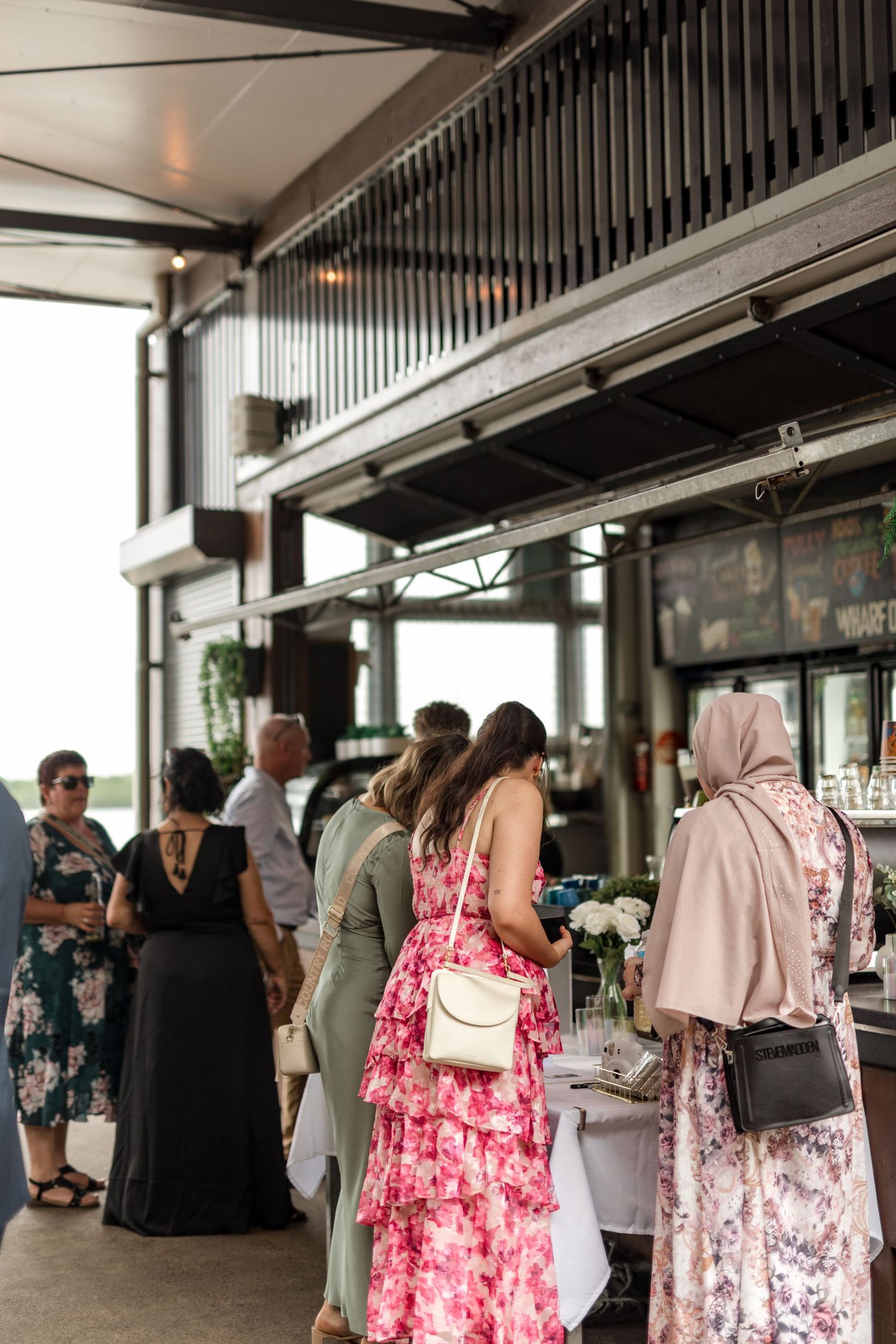 Diverse wedding guests in formal attire gathering near a bar at a modern reception venue.