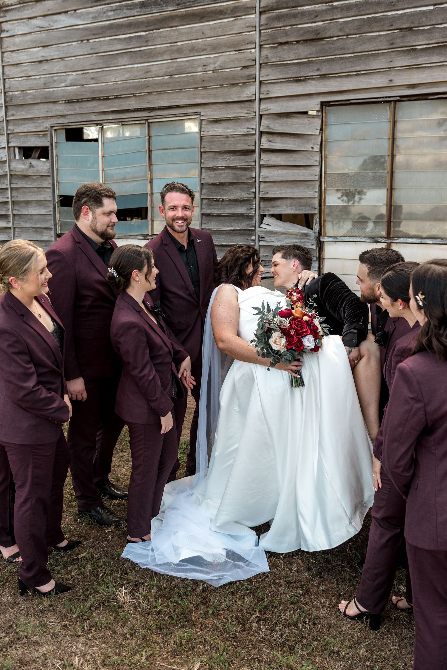 Smiling wedding party in burgundy suits surrounds the kissing bride and groom outside a weathered wooden barn.