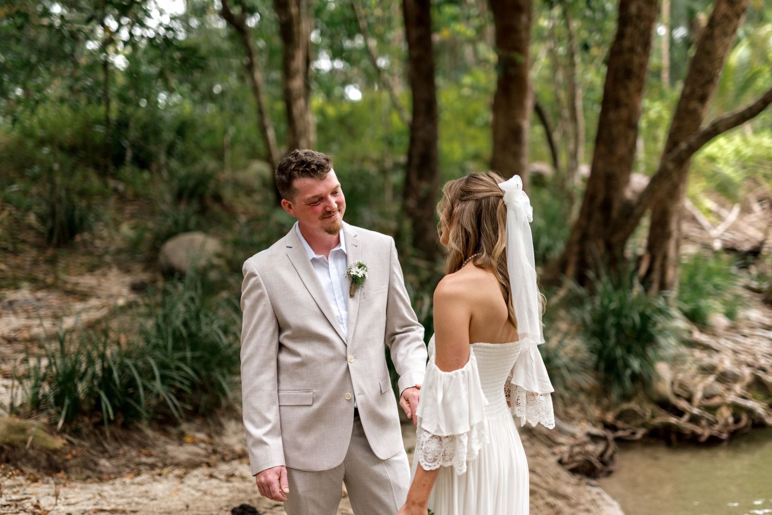 Smiling groom in beige suit turns to bride in white off-shoulder dress by river.